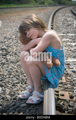 Lonely bambino siede sui binari del treno con il suo orsacchiotto Foto Stock