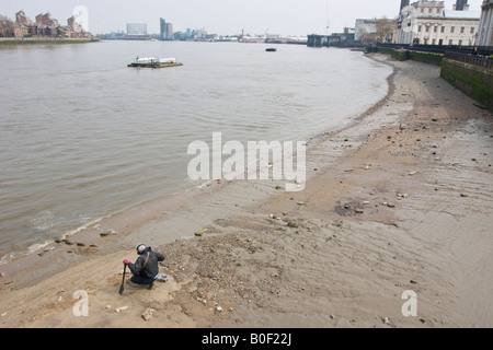 L'uomo con il rivelatore di metalli accovacciato per esaminare una ricerca sul Fiume Tamigi Foreshore a Greenwich Foto Stock