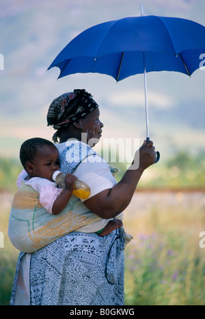 Una madre che porta un bambino sulla schiena in una imbracatura, tenendo un ombrello fino contro il sole, Sud Africa. Foto Stock