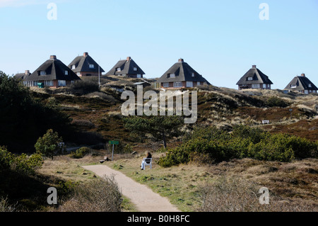 Donna seduta su una panchina accanto a un percorso che conduce a un cluster di paglia-case con tetti in Hoernum sul Nord Frisone isola di S Foto Stock