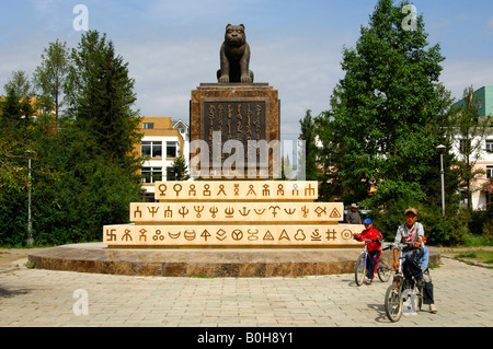 Bambini ciclismo passato il monumento alla Stato mongolo guarnizione, scultura di una tigre, iscritto nel vecchio script mongola, Ulaanbat Foto Stock