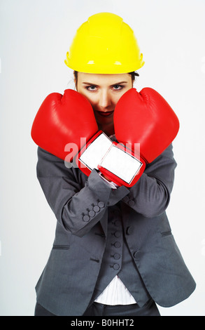 Giovane imprenditrice, manager rosso indossa guantoni da pugilato e giallo hardhat difendendo se stessa Foto Stock