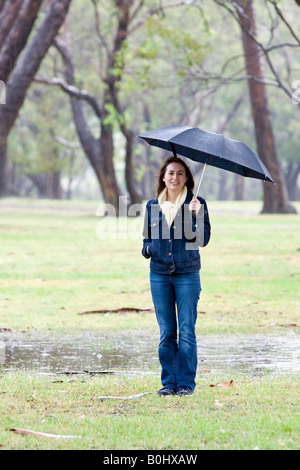 Una giovane donna con un ombrello sotto la pioggia, sorgeva accanto ad una pozza in un parco. Foto Stock