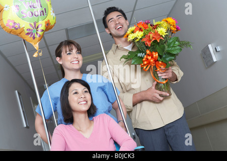 Infermiere e uomo con bouquet di assegnare una donna su una sedia a rotelle in ospedale in corridoio Foto Stock