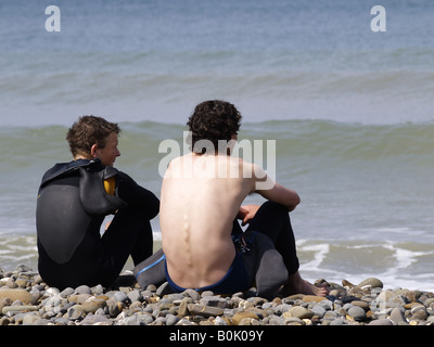 Due surfisti seduto su di una spiaggia di ciottoli, guardando le onde, Cornwall, Regno Unito Foto Stock