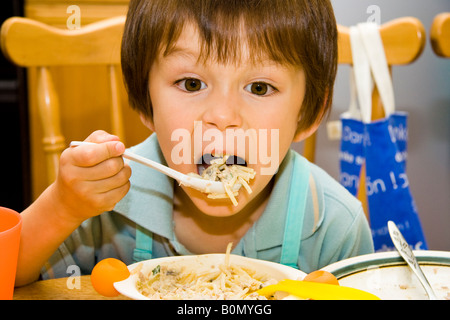 Primo piano di bambino caucasico, ragazzo di 4-5 anni, di fronte, seduto mentre solleva un folklore di plastica carico di pasta alla sua ampia bocca aperta. Foto Stock