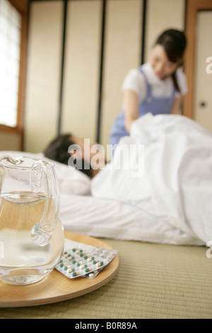 Brocca da acqua e pillole medicinali sul vassoio con il senior donna recante sul materasso e infermiere su sfondo Foto Stock