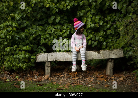 Carino quattro anno vecchia ragazza siede su legno banco registro che indossa un capo di lana bobble hat Foto Stock