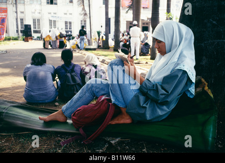 Una donna musulmana siede contro un albero e creare note attorno ad una parte in Indonesia Foto Stock