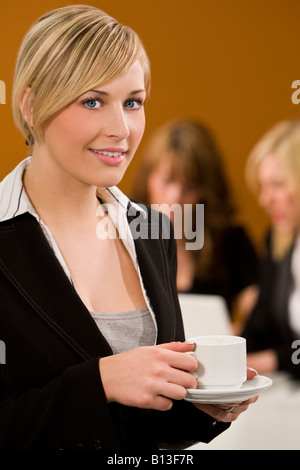 Un attraente giovane femmina executive prende una pausa caffè mentre i suoi colleghi di lavoro su un computer portatile dietro di lei Foto Stock