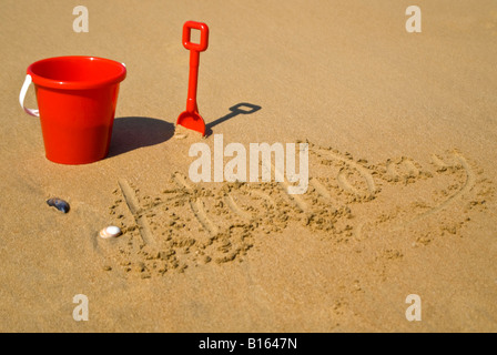 Vista orizzontale di un rosso secchio di plastica e vanga sulla spiaggia accanto a "vacanza" scritto nella sabbia Foto Stock