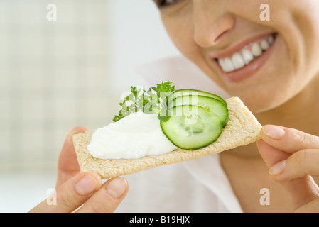 Giovane donna tenendo premuto fino cracker conditi con cetrioli e formaggio spalmabile, ritagliato Foto Stock