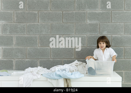 Piccolo Ragazzo seduto in contenitore in plastica, servizio lavanderia disseminata accanto a lui Foto Stock