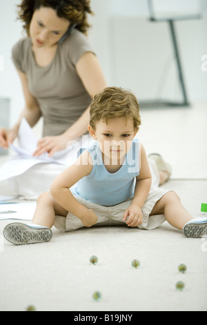 Ragazzo giocando con marmi mentre la giovane madre utilizza il telefono e guarda la documentazione in background Foto Stock