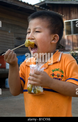 Un piccolo ragazzo tailandese mangia uno spuntino che ha appena comprato da un prodotto alimentare venditore ambulante Foto Stock