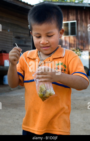 Un piccolo ragazzo tailandese mangia uno spuntino che ha appena comprato da un prodotto alimentare venditore ambulante Foto Stock