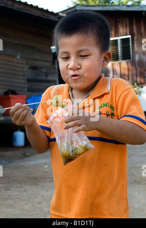 Un piccolo ragazzo tailandese mangia uno spuntino che ha appena comprato da un prodotto alimentare venditore ambulante Foto Stock