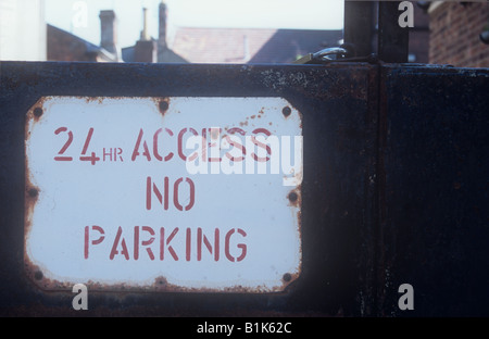 Il bianco e il rosso avviso su arrugginendo metallo solido e bloccato i cancelli dichiarando 24 hr Access non Parcheggio con tetti visibile oltre Foto Stock