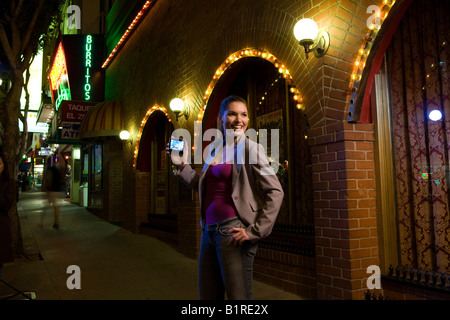 Una giovane donna si trova in una ben illuminata street al calar della sera che porta un dispositivo tenuto manualmente. Foto Stock