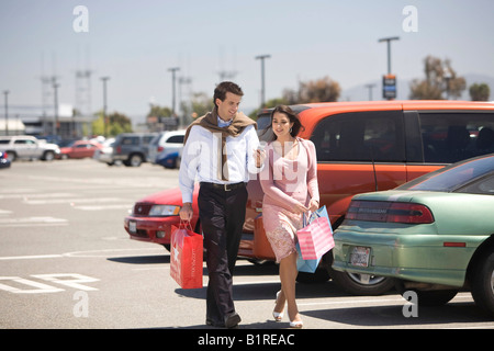 Una giovane coppia a piedi attraverso un parcheggio pieno di auto portante un dispositivo tenuto manualmente e diverse borse per lo shopping. Foto Stock