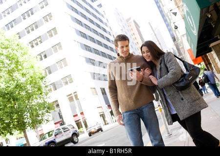 Una giovane coppia sono passeggiando sotto braccio attraverso un centro di zona della città portando un dispositivo tenuto manualmente. Foto Stock