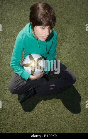 Giovane donna seduta su un campo di calcio e tenendo un calcio nelle sue braccia Foto Stock