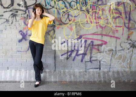 Giovane donna l'ascolto di musica con cuffie di grandi dimensioni, in piedi di fronte a graffiti parete spruzzato Foto Stock