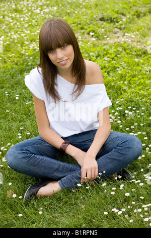 Giovani, dai capelli scuri donna che indossa jeans e un top bianco, seduti in un prato sommerly, cercando cordiale alla fotocamera Foto Stock