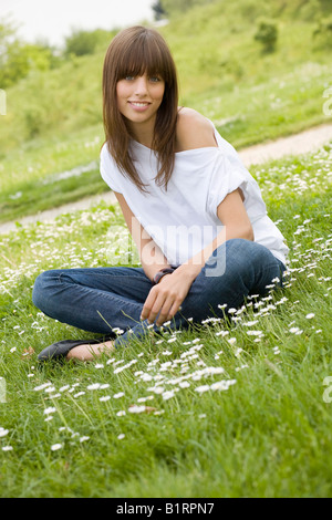 Giovani, dai capelli scuri donna che indossa jeans e un top bianco, seduti in un prato sommerly, cercando cordiale alla fotocamera Foto Stock