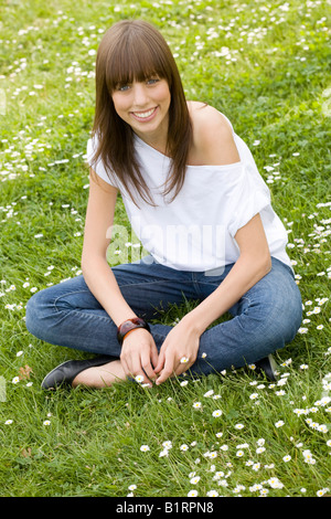 Giovani, dai capelli scuri donna che indossa jeans e un top bianco, seduti in un prato sommerly, cercando cordiale alla fotocamera Foto Stock