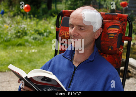 L'uomo, 45, con un bendaggio adesivo sulla sua testa seduto in una sedia da giardino leggendo un libro, Geretsried, Baviera, Germania, Europa Foto Stock