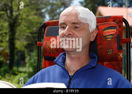 L'uomo, 45, con un bendaggio adesivo sulla sua testa seduto in una sedia da giardino leggendo un libro, Geretsried, Baviera, Germania, Europa Foto Stock