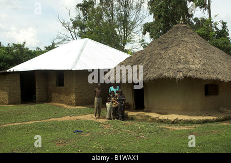 Due capanne di fango con la famiglia al di fuori di uno con tetto di paglia e uno con tin roof costruito con procedes da una carità donata mucca Foto Stock