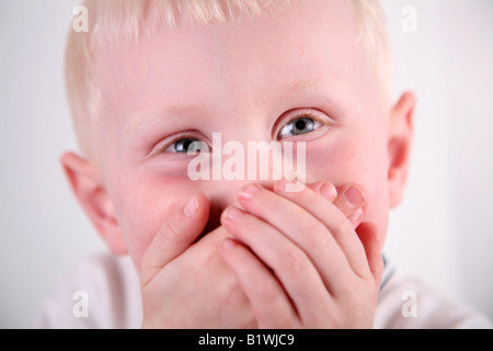 Immagine ravvicinata contro uno sfondo bianco di una giovane dai capelli biondo ragazzo che copre la sua bocca con entrambe le mani mentre sogghignando. Foto Stock