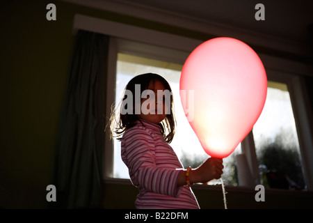 Ragazza di età compresa tra i quattro gioca con palloncino rosso illuminato dal sole del pomeriggio streaming attraverso una finestra. Razza mista asian inglese Foto Stock