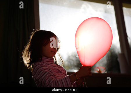 Ragazza di età compresa tra i quattro gioca con palloncino rosso illuminato dal sole del pomeriggio streaming attraverso una finestra Foto Stock