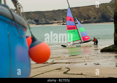 Due marinai tirare un vivacemente colorato catamarano barca a vela su una spiaggia al di fuori della parete del porto in Newquay Cornwall Inghilterra Foto Stock