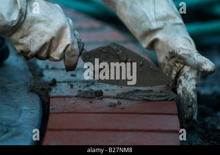 Le mani con guanti di un muratore con una cazzuola per posizionare la malta tra i mattoni di argilla rendendo il bordo di un patio del cortile in California. Foto Stock