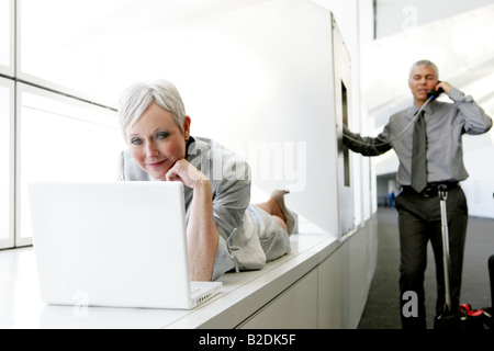 Felice donna matura utilizzando il portatile in aeroporto con Uomo al telefono in background. Foto Stock