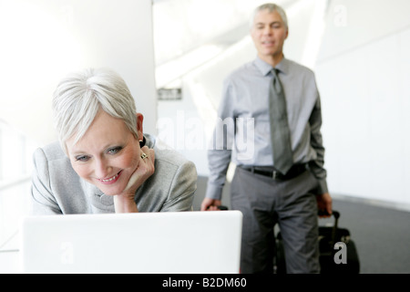 Felice donna matura utilizzando il portatile in aeroporto con uomo dietro. Foto Stock