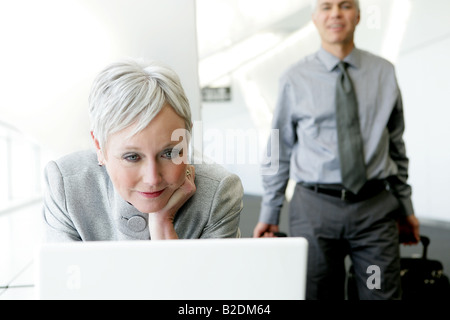 Donna matura utilizzando il portatile in aeroporto con uomo dietro. Foto Stock