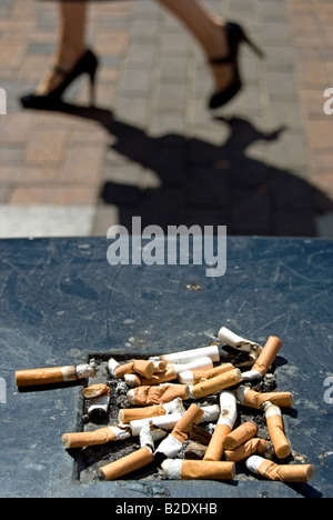 Scartato mozziconi di sigaretta su una strada britannico spazzatura con una donna a piedi passato in tacchi alti Foto Stock