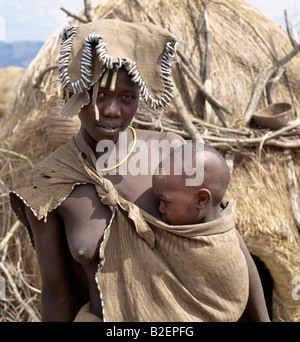 Un Mursi la madre e il bambino. La madre sfumature la sua testa con uno dei suoi grembiuli di pelle che è bordato con anelli di metallo. Foto Stock