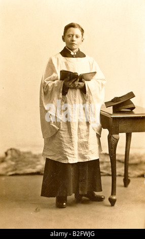 1910-1915 fotografia d'annata, ritratto in studio di un giovane uomo in piedi, 10-15 anni, che indossa un costume da coro e tiene libro di Inno. Contatto con gli occhi. Foto Stock