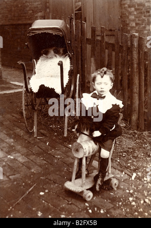 1905 Italiano seppia edoardiana Fotografia di un bambino di 3-4 anni seduto su un trolley tipo cavallo hobby in giardino. PRAM in background. Foto Stock