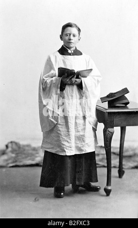 1910-1915 fotografia d'annata, ritratto in studio di un giovane uomo in piedi, 10-15 anni, che indossa un costume da coro e tiene libro di Inno. Contatto con gli occhi. Foto Stock