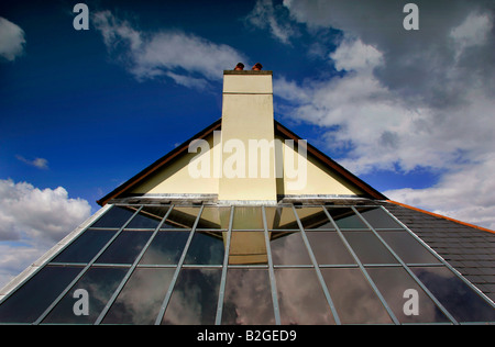 Pannelli solari su un tetto in Devon UK che riflette un tetto spiovente e il camino contro un cielo blu con nuvole. Foto Stock