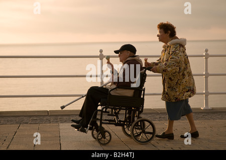 Donna anziana caregiver spingendo il suo marito di disabili in sedia a rotelle sia a mangiare il gelato mare serata estiva, Wales UK Foto Stock