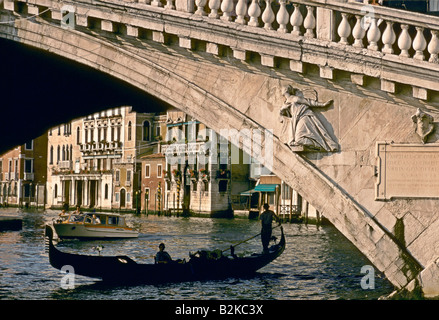 Una gondola con passeggero seduto e gondoliere permanente sulla sua punta passa sotto un ponte curvo con un angelo in rilievo su di esso Foto Stock