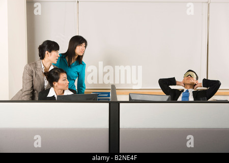 Tre imprenditrici guardando un imprenditore di dormire su una sedia in un ufficio Foto Stock